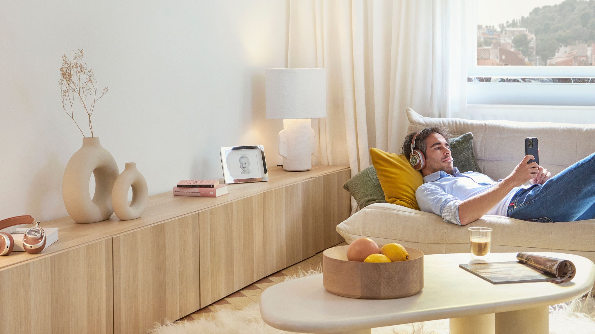 En avslappnad man som ligger på soffan i vardagsrummet och lyssnar med hörlurar och skrollar på telefonen